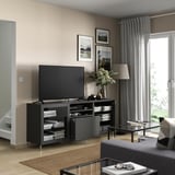 Modernes Wohnzimmer mit BESTÅ-TV-Bank, Flachbildschirm, grauen Vorhängen, Holzboden, Teppich, Lampe, Blumen, gerahmten Fotos, schwarzem Couchtisch.
