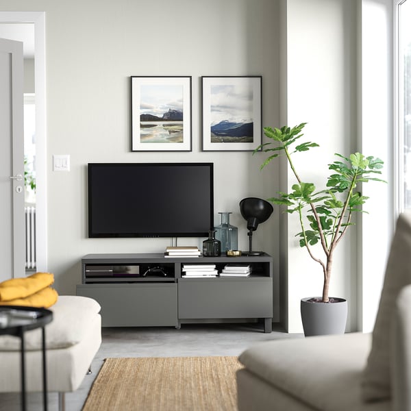 Modernes Wohnzimmer mit BESTÅ-TV, Flachbildfernseher, gerahmten Fotos, weißem Sofa, gelbem Überwurf, großer Pflanze, grauem Teppich und schwarzer Lampe.