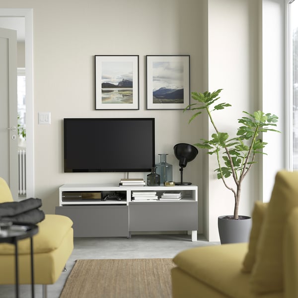 Wohnzimmer mit Wandfernseher auf grauem Ständer, mit zwei gelben Sesseln, einer großen grünen Pflanze und zwei gerahmten Bergfotos.