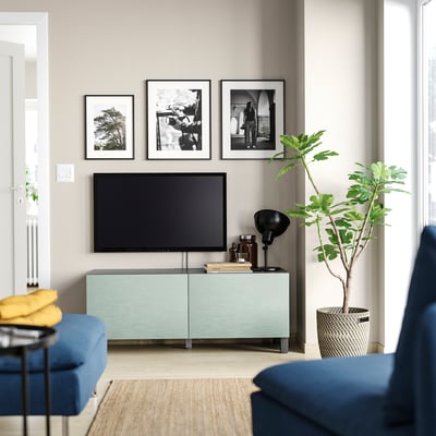 Modernes, minimalistisches Wohnzimmer mit grünem BESTÅ-TV, Flachbildfernseher, Dekor, blauen Sofas, Pflanzen und gerahmter Wandkunst.