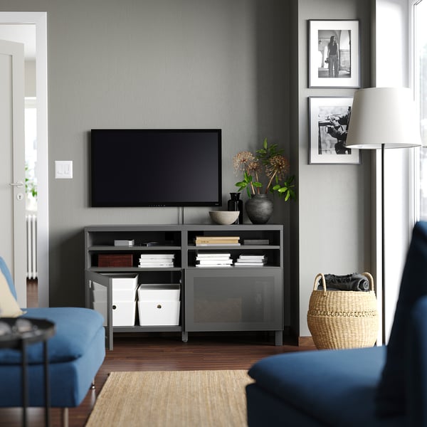 Modernes Wohnzimmer mit grauem BESTÅ-TV-Ständer, blauem Sofa, Holzboden, schwarzem Fernseher, weißer Lampe, geflochtenem Korb und Wandbildern.