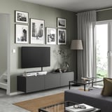 Modernes Wohnzimmer mit BESTÅ-TV-Bank in Grau, Flachbildfernseher, gerahmten Schwarz-Weiß-Fotos, grauen Vorhängen, Tischlampe und Couchtisch aus Glas.