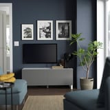 Modernes Wohnzimmer mit dunklen Wänden, grauer BESTÅ-TV-Bank, Großbildfernseher, gelbem Kissen, grüner Pflanze, viel Stauraum.