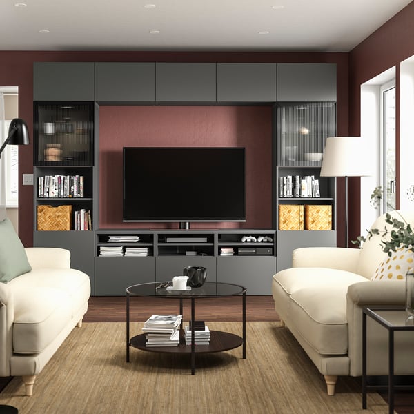Modernes Wohnzimmer mit dunkelroter Akzentwand, großem Flachbildfernseher auf grauer BESTÅ-TV-Bank, weißen Sofas, Glascouchtisch und Tageslicht.