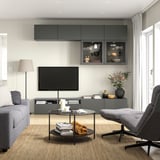 Modernes Wohnzimmer mit grauer BESTÅ-TV-Bank, Flachbildfernseher, grauem Sofa, Sessel, Glascouchtisch und Wandregalen.