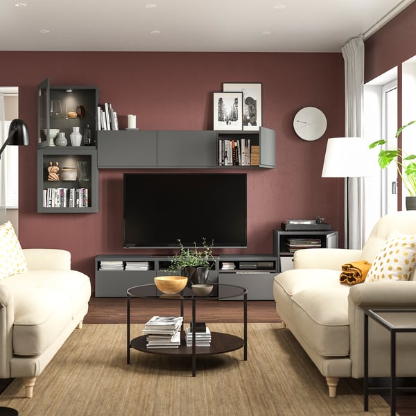 Modernes Wohnzimmer mit kastanienbraunen Wänden, schwarzer BESTÅ-TV-Bank, beigen Sofas mit gelben Kissen, Couchtisch aus Glas und Regalen.
