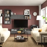 Modernes Wohnzimmer mit kastanienbraunen Wänden, schwarzer BESTÅ-TV-Bank, beigen Sofas mit gelben Kissen, Couchtisch aus Glas und Regalen.