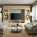 Ein gemütliches Wohnzimmer mit Fernseher, Sofas, Glastisch und Stehlampe. Helles Holz, natürliches Licht, dunkelgrüne Wände.