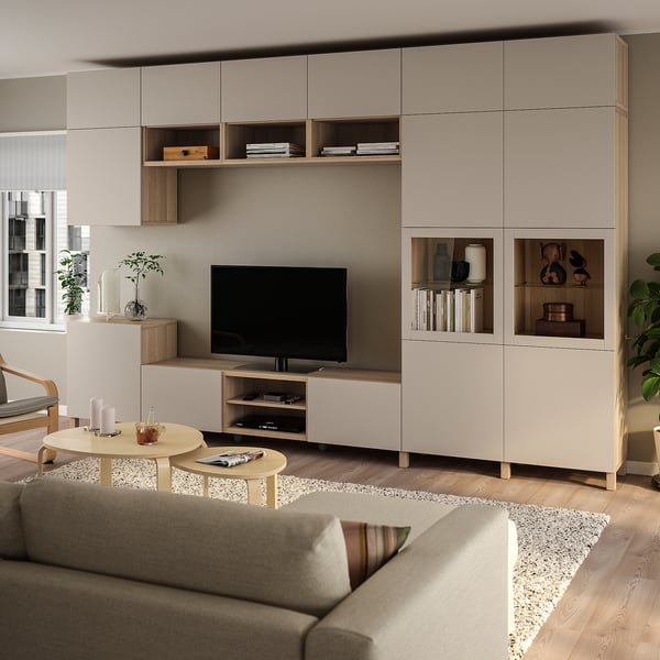 Modernes Wohnzimmer mit BESTÅ-Stauraum und TV. Neutrale Töne, graues Sofa, Holzmöbel, großer Fernseher, natürliches Licht durch Jalousien.