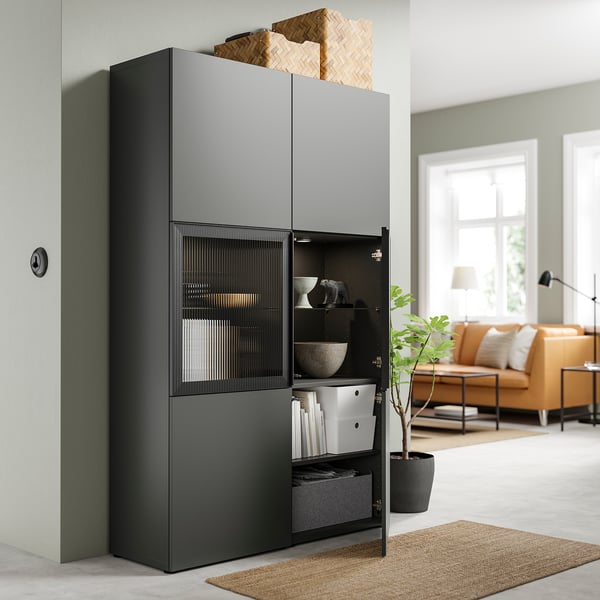 Modernes Wohnzimmer mit dunkelgrauem BESTÅ-Schrank, verstellbaren Regalen, Glastür und geflochtenen Körben obendrauf.