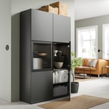 Modernes Wohnzimmer mit dunkelgrauem BESTÅ-Schrank, verstellbaren Regalen, Glastür und geflochtenen Körben obendrauf.