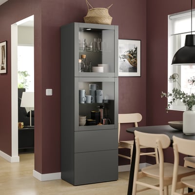 Moderner dunkelgrauer BESTÅ-Schrank mit Glastüren, in dem Tassen, Schalen und Karaffen ausgestellt sind. Essbereich aus Holz mit Stühlen und Tisch.