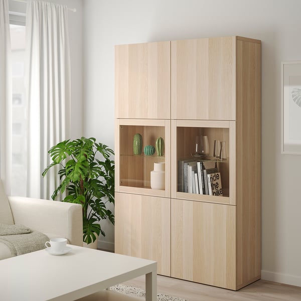 Ikea BESTÅ Aufbewahrungsschrank aus heller Eiche mit Glastüren und gemischten Regalen neben Couchtisch und Sofa.