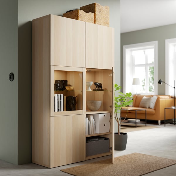 Holzschrank mit Glastüren, in dem Dekorationsgegenstände und Bücher ausgestellt sind. Eine geöffnete Tür zeigt verstellbare Regale und Aufbewahrungsboxen.