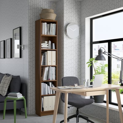 Ein gut organisiertes Homeoffice mit einem hölzernen Bücherregal, einem Bürostuhl an einem Schreibtisch am Fenster mit Laptop und Pflanze und einem gemütlichen Sofa mit Kissen.