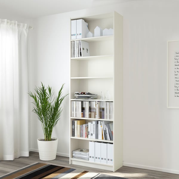 Weißes BILLY Bücherregal mit Büchern und Schachteln, in der Nähe des Fensters, grüne Pflanze, gestreifter Teppich.