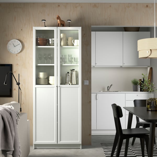 Moderne Küche mit weißem BILLY-Schrank, Glastüren, schwarzen Stühlen und Holzakzenten. Eine Uhr und Dekorationsgegenstände werten den Raum auf.