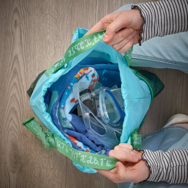 Eine Person hält ein offenes Handtuch mit blau-grünen Schildkrötenmotiven in der Hand und zeigt darin eine Schwimmmaske. Geeignet zum Trocknen und Erwärmen nach dem Baden.