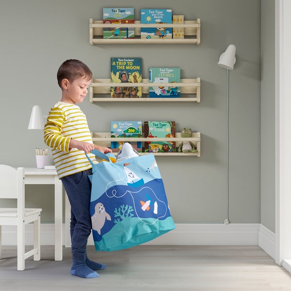 Kind in gestreiftem Hemd mit einer Aufbewahrungstasche mit Meeresmotiv. Hinter ihm stehen Regale voller bunter Kinderbücher.