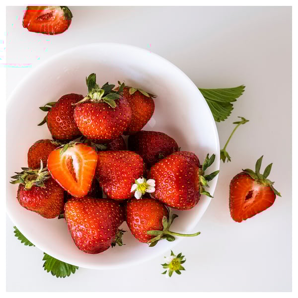Leuchtend rote Erdbeeren in weißer Schale; Einer in Scheiben geschnitten, Blätter verstreut.