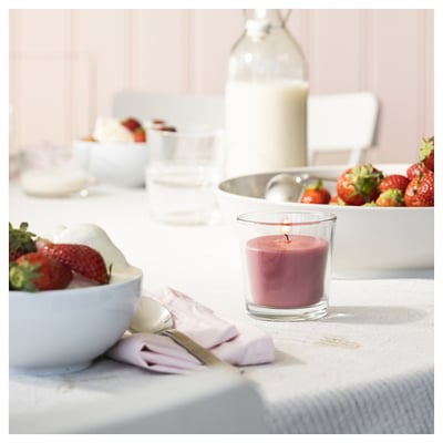 Stellen Sie sich eine brennende rosa Duftkerze in einem Glas auf einem Esstisch vor. Um ihn herum stehen Schalen mit Erdbeeren, eine Flasche und eine Serviette.