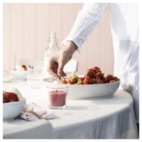 Die Person gießt rote Flüssigkeit in das Glas in der Nähe der Schüssel mit Erdbeeren auf dem Tisch.