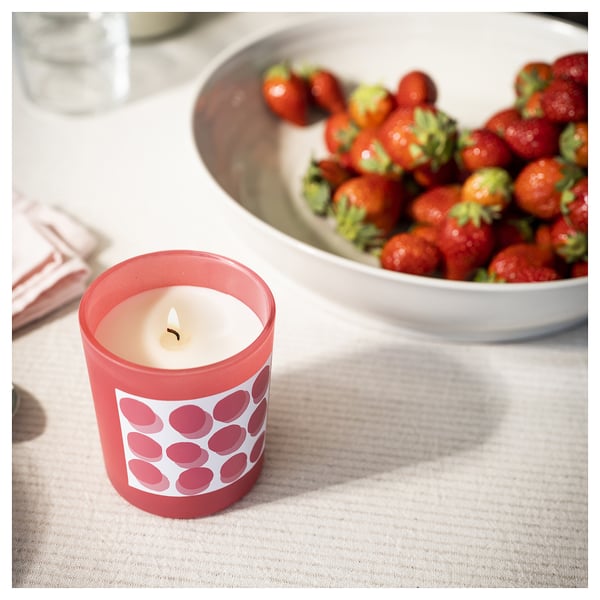 Brennende rote Kerze mit Erdbeermuster neben einer Schüssel mit frischen Erdbeeren auf weißer Tischdecke.