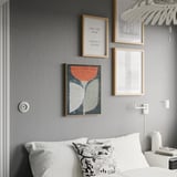 Modernes Schlafzimmer mit buntem BLODFLÄDER Poster. Drei gerahmte Drucke an grauer Wand, Bett mit weißen Kissen, Pendelleuchte.