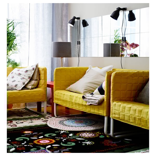 Helles Wohnzimmer mit gelben Stühlen auf einem bunten Teppich, umgeben von Spiegeln und Pflanzen.