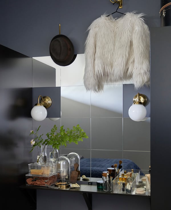 Modernes graues Badezimmer mit verspiegelter Wand, goldenen Lichtern und weißer Jacke neben dem Waschbecken. Pflanzen, Make-up, Pinselset unten.