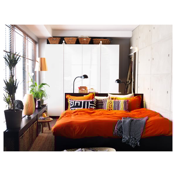 Helles Schlafzimmer mit orangefarbener Bettwäsche, gemusterten Kissen, weißen Schränken und einem schwarzen Beistelltisch mit grünen Pflanzen und einer hohen Lampe, die für Beleuchtung sorgt.