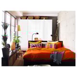 Helles Schlafzimmer mit orangefarbener Bettwäsche, gemusterten Kissen, weißen Schränken und einem schwarzen Beistelltisch mit grünen Pflanzen und einer hohen Lampe, die für Beleuchtung sorgt.