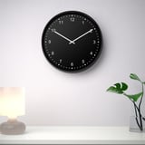 Eine moderne, runde, schwarze, lautlose Wanduhr mit weißen Ziffern und Zeigern auf 10:10 Uhr. Er ist über einem weißen Regal mit einer Lampe und einer Pflanze montiert.