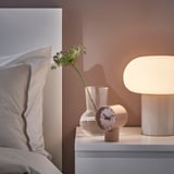 Uhr, Lampe und grüne Blumen auf dem Nachttisch. Alternativ gemütlicher Nachttisch mit Uhr, Lampe und grünen Blumen.