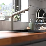 Moderne Küchenspüle mit BOSJÖN-Armatur aus Edelstahl, Seifenspender und Geschirrkorb. Der Wasserhahn steht hoch zwischen den Werkzeugen, in der Nähe eines gefliesten Spritzschutzes.