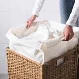 Person, die weiße Wäsche aus einem geflochtenen Rattan-Wäschekorb mit Liner-Beutel hebt.