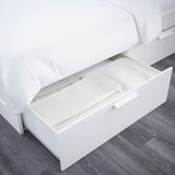 Offenes weißes Bett mit Schublade, in der gefaltete Bettwäsche zu sehen ist. Modernes, platzsparendes Design.