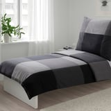 Gepflegtes Bett mit BRUNKRISSLA grau/schwarzer Bettdecke, kleines Zimmer.