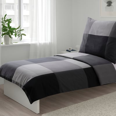 Gepflegtes Bett mit BRUNKRISSLA grau/schwarzer Bettdecke, kleines Zimmer.