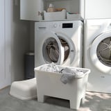 Weiße Plastikhocker und Waschmaschinen in der Waschküche. Auf dem zentralen Hocker kann die Wäsche aufbewahrt werden.