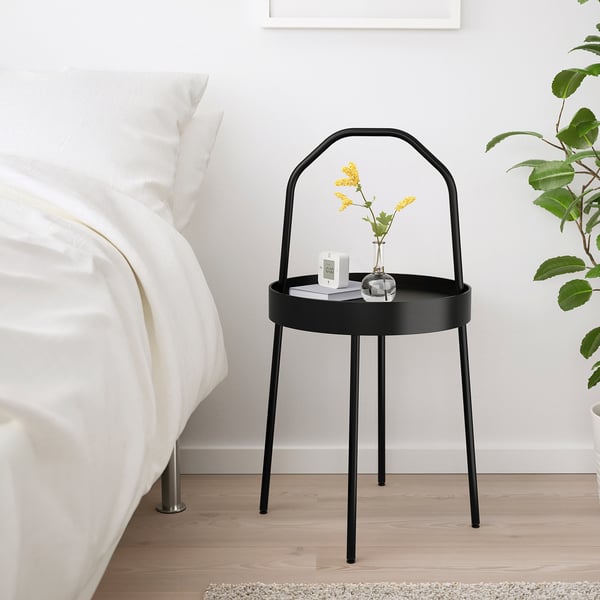 Moderner, minimalistischer Nachttisch aus schwarzem Metall neben dem Bett, mit kleiner Vase und Wecker, neben großer grüner Pflanze.