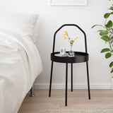 Moderner, minimalistischer Nachttisch aus schwarzem Metall neben dem Bett, mit kleiner Vase und Wecker, neben großer grüner Pflanze.