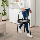 Person hält tragbaren, runden Beistelltisch mit Griff. Auf dem Tisch stehen zwei Gläser und Untersetzer. Der Tisch hat ein schwarzes Metallgestell und eine helle Platte.