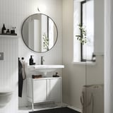 Modernes Badezimmer mit weißem Byasjøn-Waschtisch. Runder Spiegel oben, kleines Waschbecken, schwarze Accessoires. Weißer Schrank unter der Spüle, kleine Pflanze auf dem Fensterbrett.