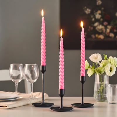 Tisch gedeckt mit leuchtenden rosa gedrehten Kerzen in schwarzen Haltern, Gläsern, Tellern und einer Vase mit weißen Blumen.