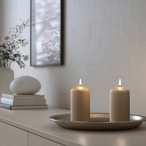 Zwei brennende beige DAGLIGEN-Kerzen auf einem Metalltablett, kleiner als Bücher und eine Vase mit Blumen, elegantes Dekor.