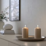 Zwei brennende beige DAGLIGEN-Kerzen auf einem Metalltablett, kleiner als Bücher und eine Vase mit Blumen, elegantes Dekor.