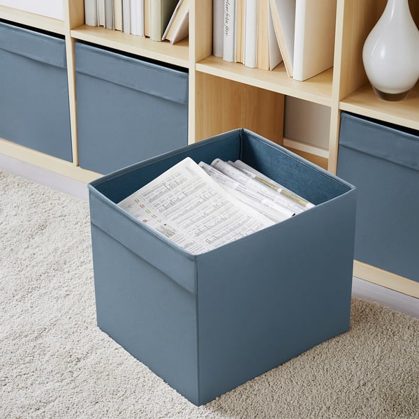 Blaue DRÖNA Aufbewahrungsbox mit Papieren auf beigem Teppich, kleiner als nahegelegenes Bücherregal und dekorative Vase.