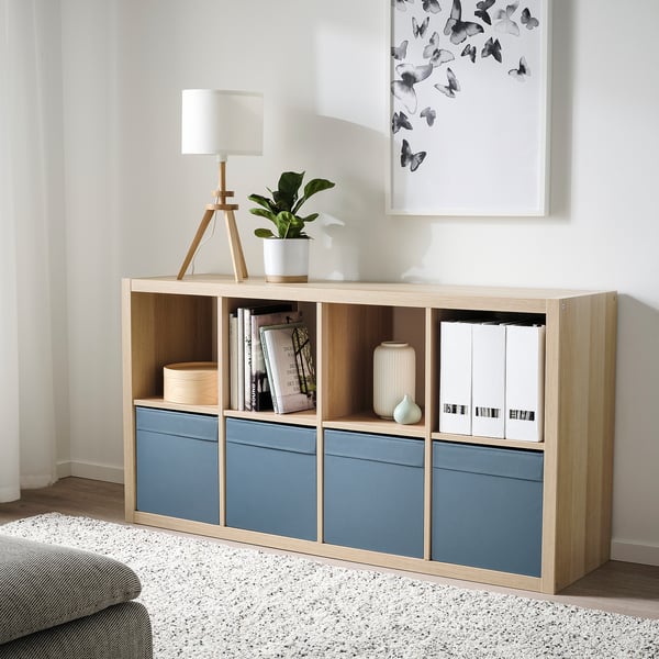 Minimalistischer Wohnraum mit hellem Holz und blauem Stauraum. Er hält Bücher, eine Lampe und Pflanzen. Die Schachtel ist schmal und passt unter ein Kunstwerk.