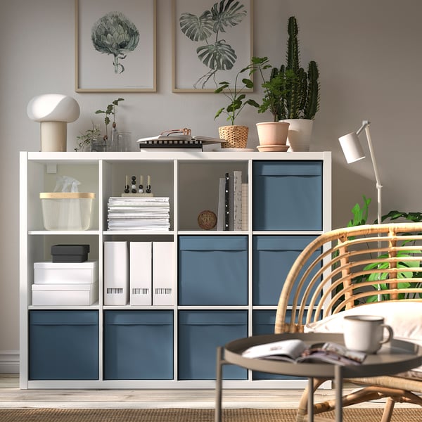 Moderner Wohnraum mit blauem Stauraum, Büchern, Pflanzen und Dekor. Korbsessel mit grünen Kissen im Vordergrund.